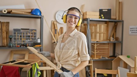 Photo for Smiling woman with safety gear holding lumber in a carpentry workshop - Royalty Free Image