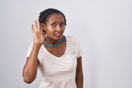 Foto de Mujer africana con rastas de pie sobre fondo blanco sonriendo con la mano sobre el oído escuchando un rumor o chismes. concepto de sordera. - Imagen libre de derechos