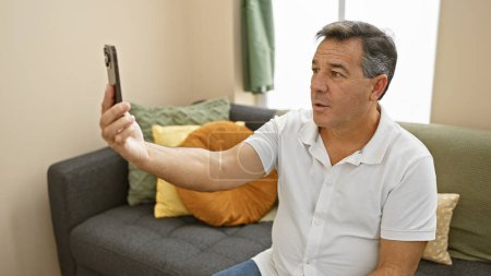 Téléchargez les photos : Un homme d'âge moyen en chemise blanche décontractée prend un selfie avec son téléphone dans un salon moderne. - en image libre de droit