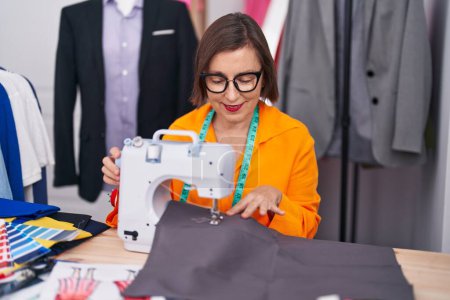 Téléchargez les photos : Femme d'âge moyen tailleur souriant confiant en utilisant une machine à coudre au magasin de tailleur - en image libre de droit