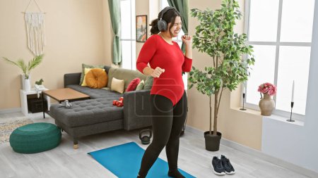Téléchargez les photos : Jeune femme hispanique dansant joyeusement dans un salon intérieur moderne tout en écoutant de la musique à l'aide d'écouteurs. - en image libre de droit