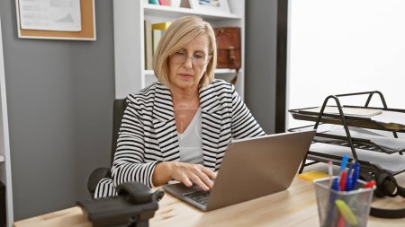 Téléchargez les photos : Femme blonde mature travaillant attentivement sur ordinateur portable dans l'intérieur du bureau moderne - en image libre de droit