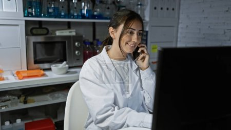Foto de Mujer hispana sonriente con bata de laboratorio hablando por teléfono en un entorno de laboratorio - Imagen libre de derechos
