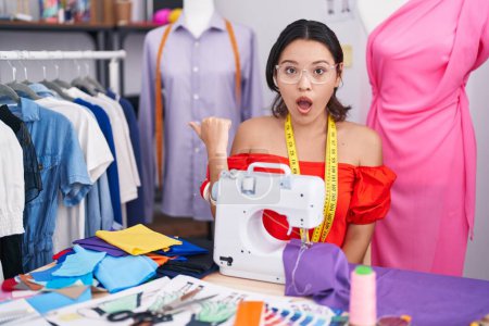Photo for Hispanic young woman dressmaker designer using sewing machine surprised pointing with hand finger to the side, open mouth amazed expression. - Royalty Free Image