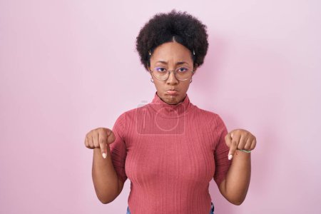 Téléchargez les photos : Belle femme africaine avec les cheveux bouclés debout sur fond rose pointant vers le bas regardant triste et contrarié, indiquant la direction avec les doigts, malheureux et déprimé. - en image libre de droit
