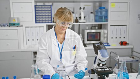 Photo for Serious, blonde middle-age woman scientist, taking notes and holding a test tube in a bustling lab, immersed in groundbreaking medical research - Royalty Free Image