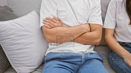 Un hombre y una mujer en atuendo casual sentados aparte en un sofá, lo que sugiere tensión en su relación en una sala de estar de tonos neutros.