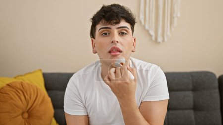 A handsome young man applies makeup with a brush in a modern living room, showcasing gender fluidity and self-care at home.
