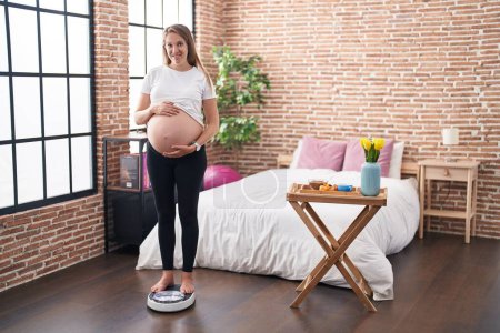 Téléchargez les photos : Jeune femme enceinte debout sur la machine de pesage toucher le ventre à la chambre - en image libre de droit