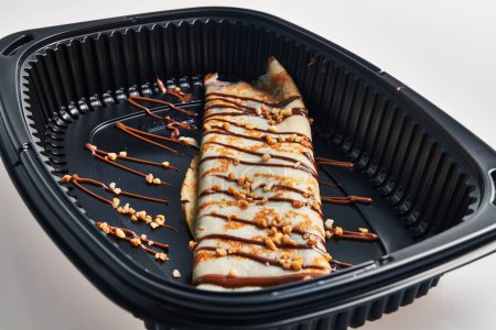 Photo for Delicious crepe adorned with chocolate and caramel drizzle in a black takeout container - Royalty Free Image