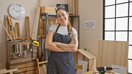 Foto de Mujer segura en delantal de mezclilla se levanta brazos cruzados en un taller de carpintería bien equipado. - Imagen libre de derechos