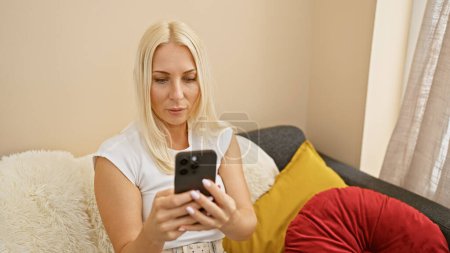 Photo for Relaxed yet serious, a beautiful blonde woman sits at home, engrossed in her online world, texting away on her smartphone in the living room. in her cozy apartment, technology meets solitude. - Royalty Free Image
