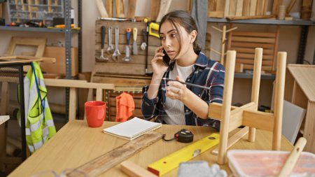 Photo for Hispanic woman in a carpentry workshop talking on phone surrounded by woodworking tools - Royalty Free Image