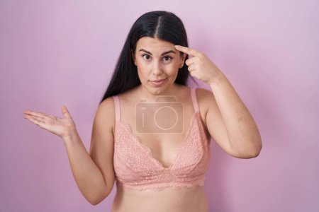 Young hispanic woman wearing pink bra confused and annoyed with open palm showing copy space and pointing finger to forehead. think about it. 