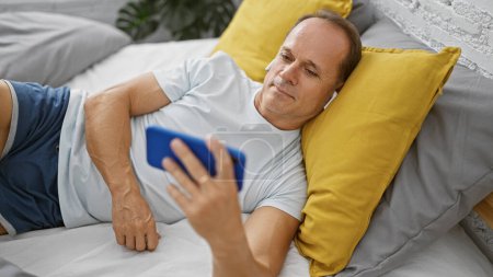 Sonriente hombre de mediana edad acostado en la cama, despierto, absorto en ver un video en su teléfono inteligente en su acogedor dormitorio