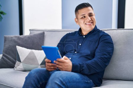 Téléchargez les photos : Jeune homme latino utilisant touchpad assis sur le canapé à la maison - en image libre de droit