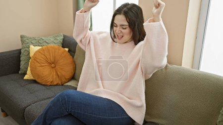 Foto de Mujer joven celebrando alegremente sola en un sofá en un acogedor salón interior, que encarna la felicidad y la positividad. - Imagen libre de derechos
