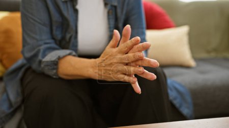 Photo for A mature hispanic woman sitting thoughtfully on a couch indoors, gesturing with her hands. - Royalty Free Image