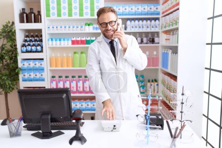 Photo for Middle age man pharmacist talking on telephone using computer at pharmacy - Royalty Free Image