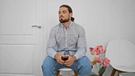 Téléchargez les photos : Jeune homme hispanique utilisant smartphone assis sur la chaise regardant bouleversé à la salle d'attente - en image libre de droit