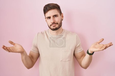 Foto de Hombre hispano con barba de pie sobre fondo rosa expresión despistada y confusa con los brazos y las manos levantadas. concepto de duda. - Imagen libre de derechos