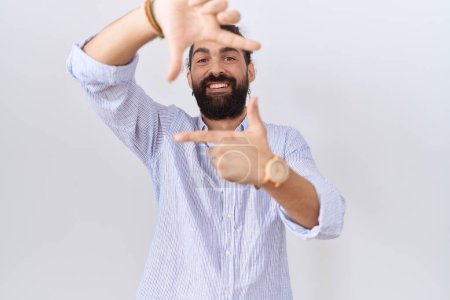 Foto de Hombre hispano con barba vistiendo camisa casual sonriente haciendo marco con manos y dedos con cara feliz. concepto de creatividad y fotografía. - Imagen libre de derechos