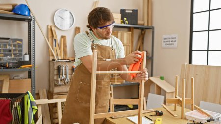 Foto de Un joven enfocado con una barba con gafas de seguridad y un delantal pule una silla de madera en un taller de carpintería bien equipado. - Imagen libre de derechos