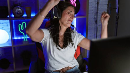 A middle-aged woman enjoys dancing while gaming in a vibrant home room at night, showcasing joy and entertainment.