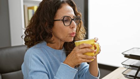Photo for Middle-aged hispanic woman with curly hair enjoying a coffee break at her modern office desk, wearing glasses and a blue sweater. - Royalty Free Image