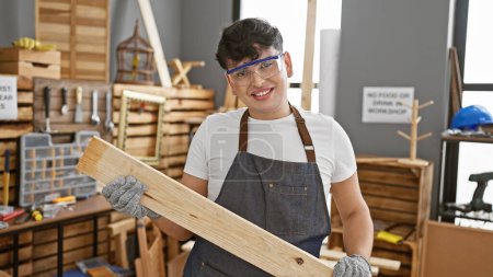 Photo for A smiling young man wearing safety glasses and an apron holds a piece of wood in a well-equipped carpentry workshop. - Royalty Free Image