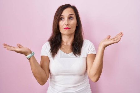 Photo for Middle age brunette woman standing over pink background clueless and confused expression with arms and hands raised. doubt concept. - Royalty Free Image