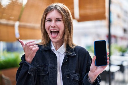 Téléchargez les photos : Joyeux jeune femme blonde tenant smartphone, rayonnant de joie tout en se tenant debout et pointant le pouce vers le côté sur la terrasse du café! - en image libre de droit