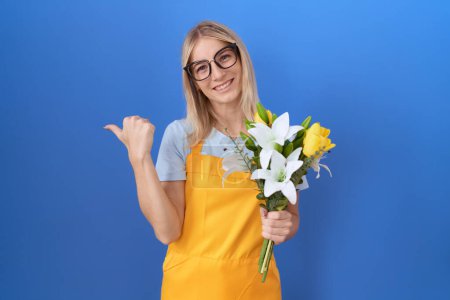 Young caucasian woman wearing florist apron holding flowers pointing to the back behind with hand and thumbs up, smiling confident 