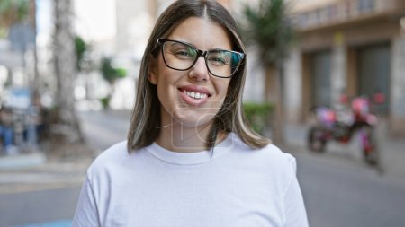 Photo for Smiling young hispanic woman wearing glasses stands on a city street, embodying urban beauty with her casual style. - Royalty Free Image
