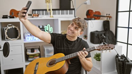 Photo for Smiling young hispanic man rocking the music studio, skillfully playing a classical guitar and making a selfie on his smartphone - Royalty Free Image