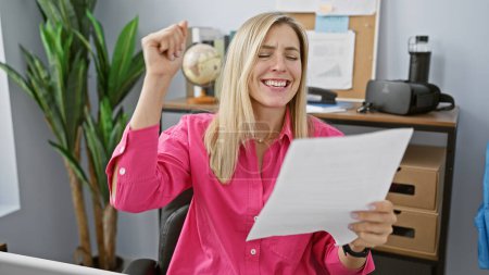 Photo for A joyful blonde woman celebrates success while reading a document in a modern office setting - Royalty Free Image