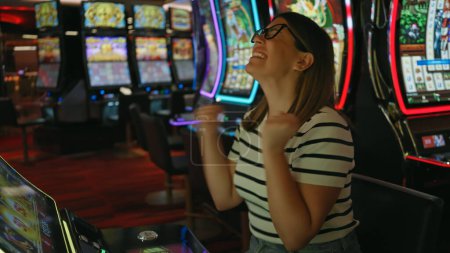 Photo for A joyful young adult woman celebrates winning at a casino slot machine. - Royalty Free Image
