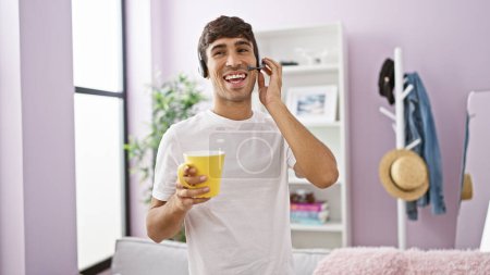 Téléchargez les photos : Souriant jeune homme hispanique heureux télétravail à la maison agent pour un centre d'appels, gadget d'appel mains libres sur, savourant du café dans le salon - en image libre de droit