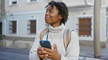 Foto de Sonriente joven mujer negra con rastas usando teléfono inteligente en una calle de la ciudad. - Imagen libre de derechos