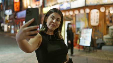 Foto de Mujer hispana segura y hermosa sonríe, tomando una selfie en la calle Tokyo, disfrutando de las luces de la ciudad por la noche, capturando la diversión a través de su teléfono inteligente. - Imagen libre de derechos