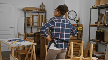 Rear-view of an adult african american woman with curly hair at her carpentry studio workshop indoors, facing shelves and a work table.