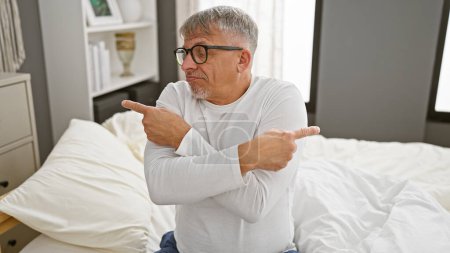 Téléchargez les photos : Un homme aux cheveux gris assis sur un lit dans une chambre bien éclairée, regardant perplexe et pointant dans deux directions différentes. - en image libre de droit
