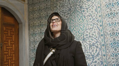 A smiling woman with glasses wearing a headscarf stands in front of decorative ottoman tiles in istanbul.