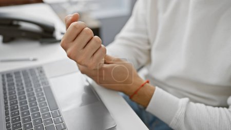 Foto de Un hombre joven adulto con barba experimenta dolor de muñeca mientras usa un portátil en un entorno de oficina. - Imagen libre de derechos