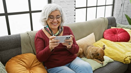 Téléchargez les photos : Une femme aux cheveux gris souriante aime utiliser un smartphone assis sur un canapé à côté de son chien au repos dans un salon confortable. - en image libre de droit