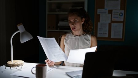 Téléchargez les photos : Une jeune femme concentrée travaille tard à son bureau, éclairée par une lampe de bureau au milieu de la paperasserie, suggérant un cadre intérieur professionnel. - en image libre de droit