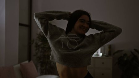 Photo for Smiling young hispanic woman stretching in her dark bedroom, portraying comfort and relaxation at home. - Royalty Free Image