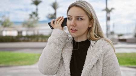 Photo for Caucasian woman in a fluffy jacket listening to a message on her smartphone outdoors. - Royalty Free Image