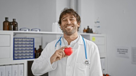 Lächelnder Mann in weißem Laborkittel mit rotem Herz in Krankenhausatmosphäre, der das Gesundheitskonzept vermittelt.