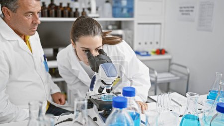 Hispanic man and woman as focused science partners, seriously working together in lab! zoom on chemistry experiment with microscope in bustling laboratory for medical research.